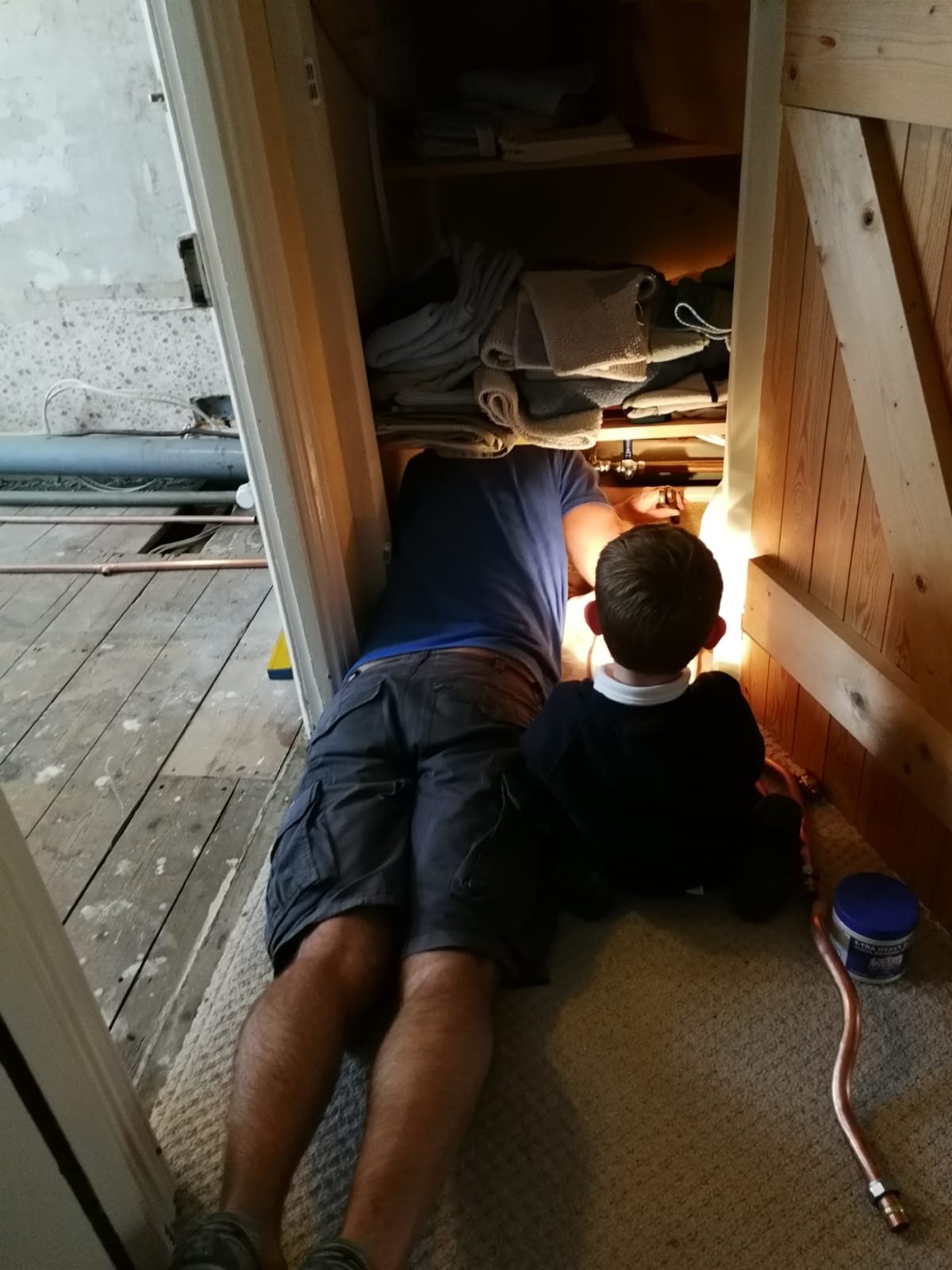 Man lying down repairing pipe in cupboard, whilst grandson holds the torch